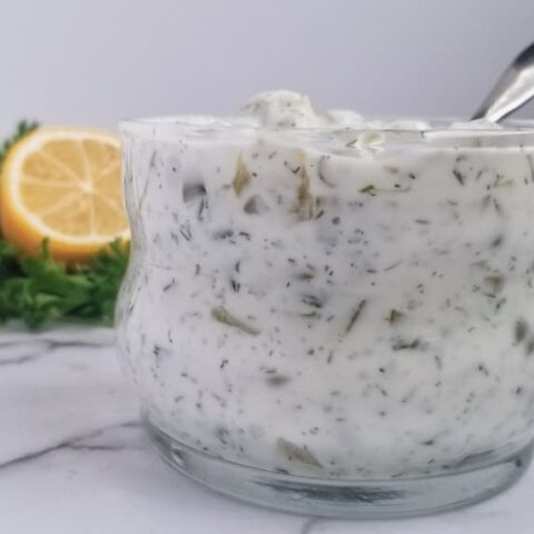 close up of a jar of quick tartar sauce sitting on a white platform, you could see pickles and fresh herbs in it, spoon in the jar, half a lemon and parsley in the background