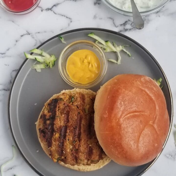 a grilled zucchini turkey burger on a bun on a plate with a small ramekin of mustard, ketchup and white dip in the background, shredded zucchini pieces around