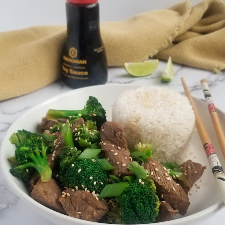 bowl of beef and broccoli, chopsticks and pile of rice in the bowl, 2 lime wedges and bottle of soy sauce in the background with a yellow tea towel