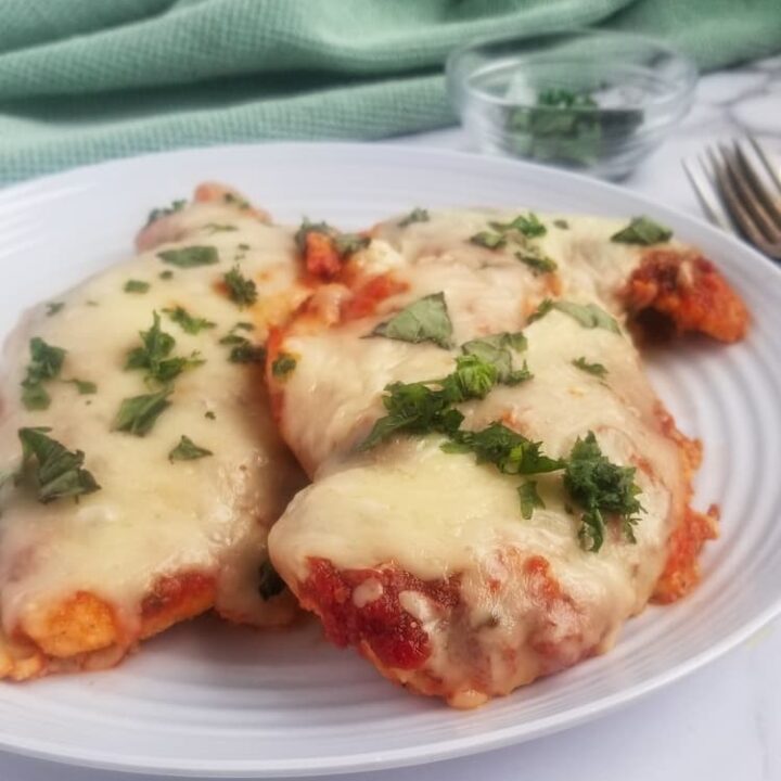 side view of plate with two cooked chicken breasts topping with mozzarella cheese and chopped fresh parsley, bowl of chopped parsley in the background. Fork and spoon on the side