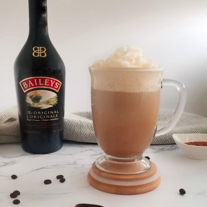 bottle of baileys next to a clear mug of irish coffee with baileys topped with whipped cream, wooden spoon and coffee beans in the front