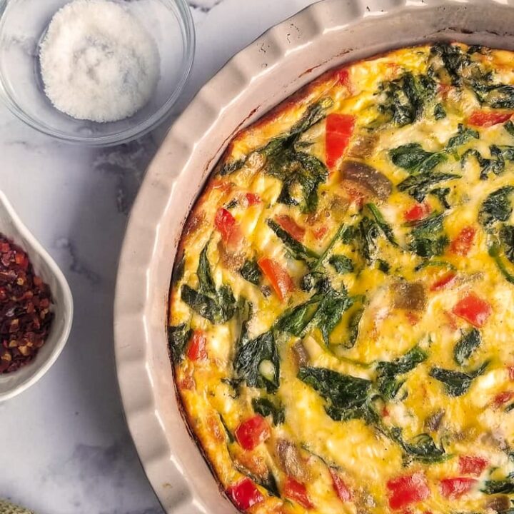 half photo of baked crustless quiche in a pie dish with spinach and red pepper, forks/plates in background with ramekins of salt and chili flakes