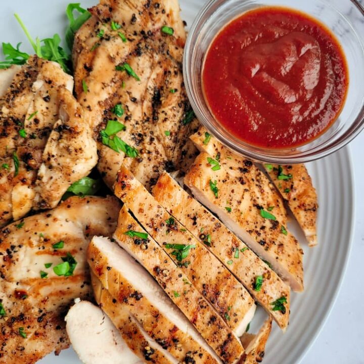 sliced grilled chicken on a plate, garnished with fresh chopped parsley, ramekin of red sauce on the plate