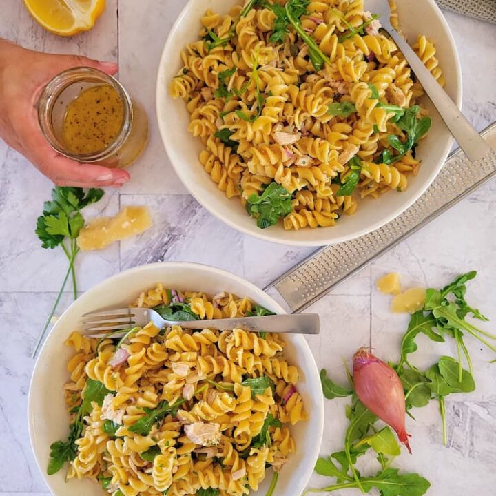 two bowls of pasta salad with tuna, arugula on the top and side with a shallot, a half a lemon and a hand holding a small jar of the dressing