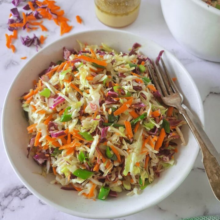 bowl of coleslaw with 2 forks, jar of dressing with a whole lime and shredded red cabbage and carrots in the background