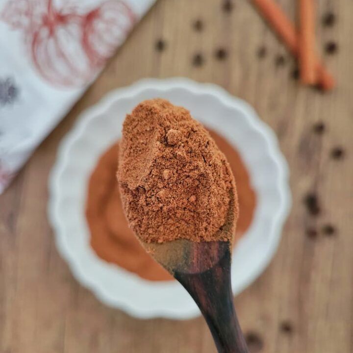 spoonful of pumpkin spice held up in front of camera lens, bowl of the rest in the background with cinnamon sticks and whole allspice