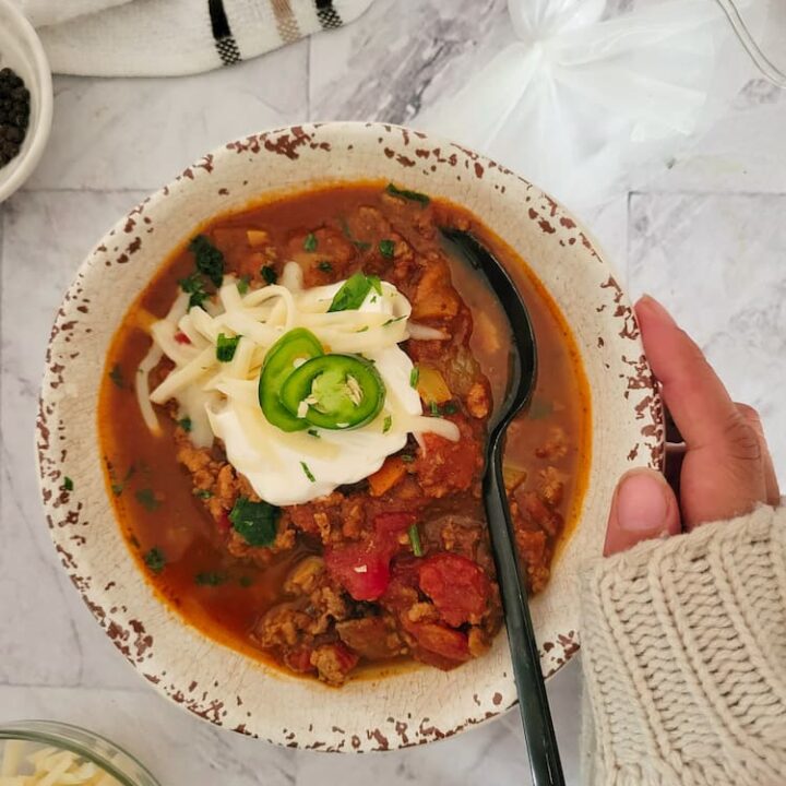 hand on a bowl of pumpkin chili with a spoon, topped with sour cream, sliced jalapeno, shredded mozzarella cheese, bowl of more cheese in the bottom background