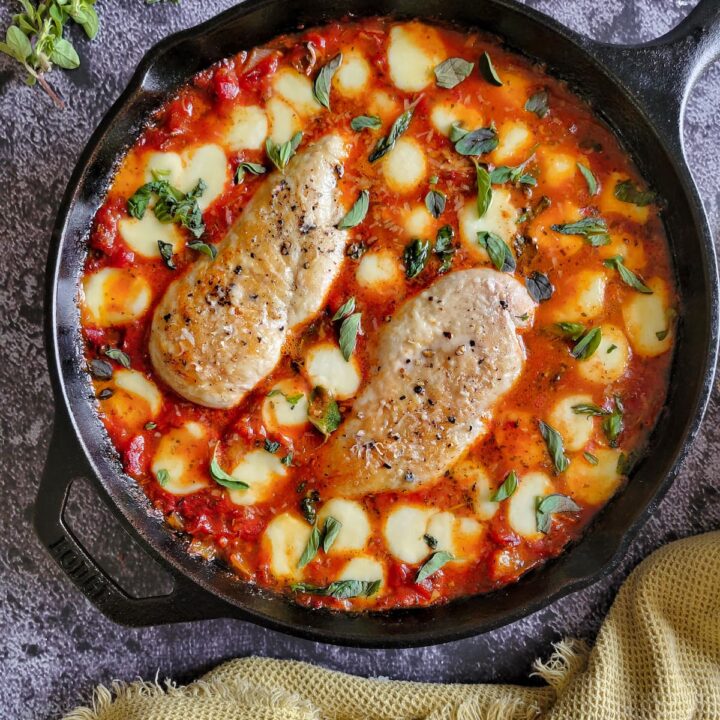 two cooked and seasoned chicken breasts in tomato sauce surrounded by melted bocconcini and garnished with chopped fresh basil in a cast iron skillet, fresh herbs in the background
