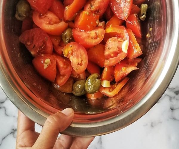 A big silver bowl contains a tomato salad - huge chunks of fresh, juicy tomatoes, chopped garlic, green pitted olives, herbs and spices, and extra virgin olive oil, a spoon sticks out the side of the bowl, hand holding bowl