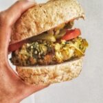 A hand holding a vegetarian burger - a spinach and artichoke millet burger patty, sandwiched between a whole wheat bun with some pickles and sliced tomato