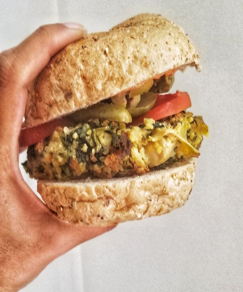 A hand holding a vegetarian burger - a spinach and artichoke millet burger patty, sandwiched between a whole wheat bun with some pickles and sliced tomato