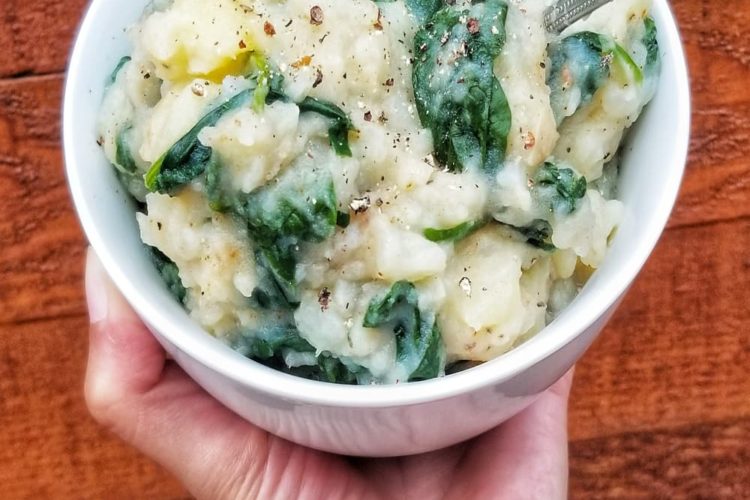 hand holding a big bowl of mashed potatoes with bright green spinach stirred into it, spoon sticking out of the side, lots of salt, pepper and chilli flakes on top