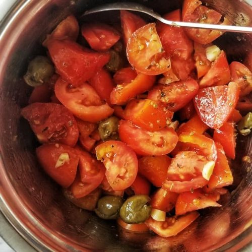 A big silver bowl contains a tomato salad - huge chunks of fresh, juicy tomatoes, chopped garlic, green pitted olives, herbs and spices, and extra virgin olive oil, a spoon sticks out the side of the bowl