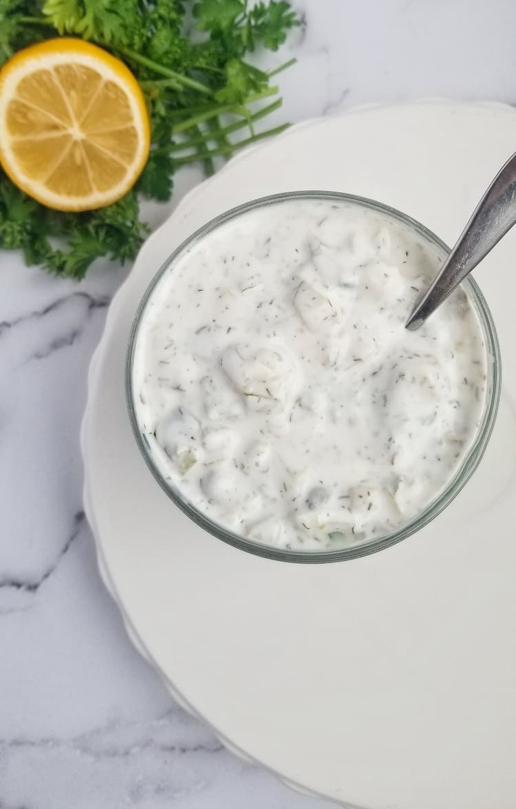 a jar of homemade tartar sauce with a spoon in it, half lemon and bunch of parsley in the backgroud