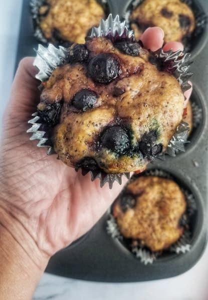 hand holding up a large freshly baked blueberry lemon poppy seed muffin over a muffin tin with the rest of the muffins