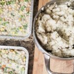 two small square trays of vegan shepherd's pie made up of white beans, broccoli, peas, corn, carrots and green beans next to a large pot of creamy potatoes