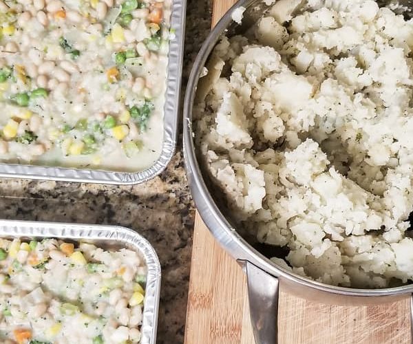 two small square trays of vegan shepherd's pie made up of white beans, broccoli, peas, corn, carrots and green beans next to a large pot of creamy potatoes