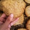 hand holding up one almond pulp cookie over a dozen more cookies on a wire rack