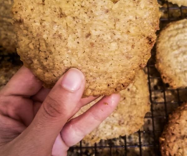 hand holding up one almond pulp cookie over a dozen more cookies on a wire rack