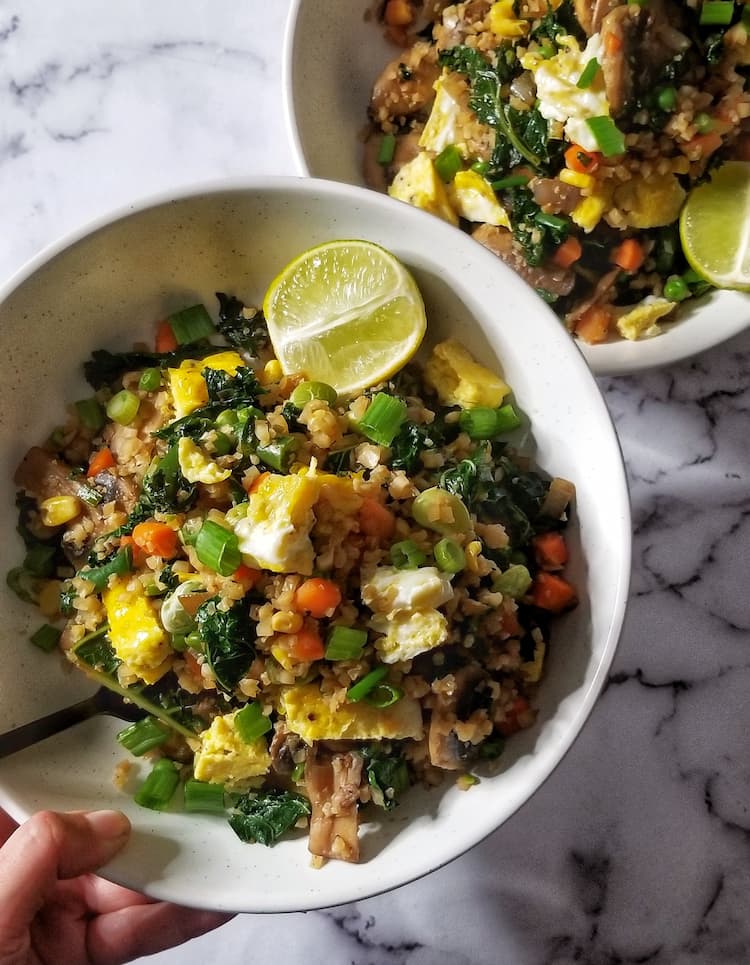 Hand holding up a delicious and colourful bowl of veggie packed cauliflower fried "rice". The dish has kale, mushrooms, frozen mixed vegetables, green onions and scrambled eggs over another bowl of the same thing. Half of a lime on each bowl