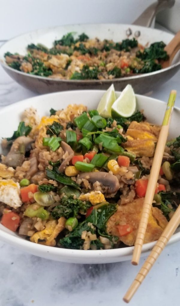 Delicious and colourful bowl of veggie packed cauliflower fried "rice". The dish has green onions, kale, mushrooms, frozen mixed vegetables and scrambled eggs, 2 lime wedges, chopsticks on top and the skillet it came out of behind it