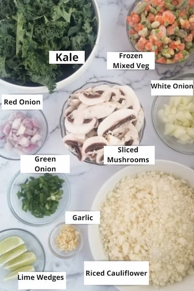some of the ingredients to make cauliflower fried 'rice' - riced cauliflower, frozen mixed veggies, kale, red onion, white onion, green onion, garlic and lime wedges