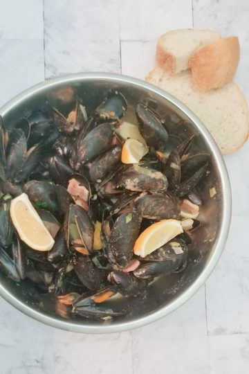 big bowl of cooked mussels with parsley, bacon, potatoes, lemon and beer broth. two slices of fresh cut bread are beside the bowl