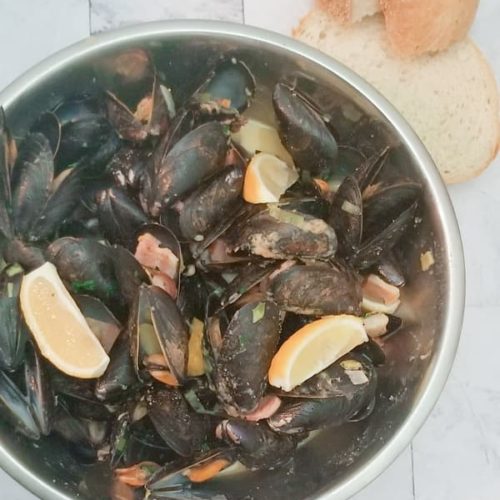 big bowl of cooked mussels with parsley, bacon, potatoes, lemon and beer broth. two slices of fresh cut bread are beside the bowl