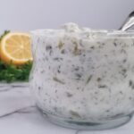 close up of a jar of quick tartar sauce sitting on a white platform, you could see pickles and fresh herbs in it, spoon in the jar, half a lemon and parsley in the background