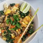 Delicious and colourful bowl of veggie packed cauliflower fried "rice". The dish has green onions, kale, mushrooms, frozen mixed vegetables and scrambled eggs, chopsticks on top, 2 lime wedges and the pan it came out of behind it
