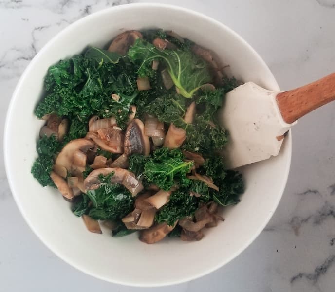 bowl of sauteed kale, mushrooms and onions with a rubber spatula in it on the side