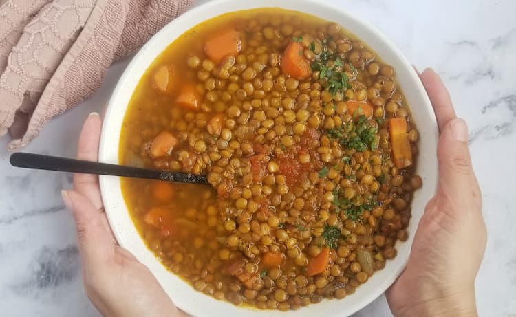 hands holding big bowl of lentil soup with carrots, tomatoes, onions and parsley