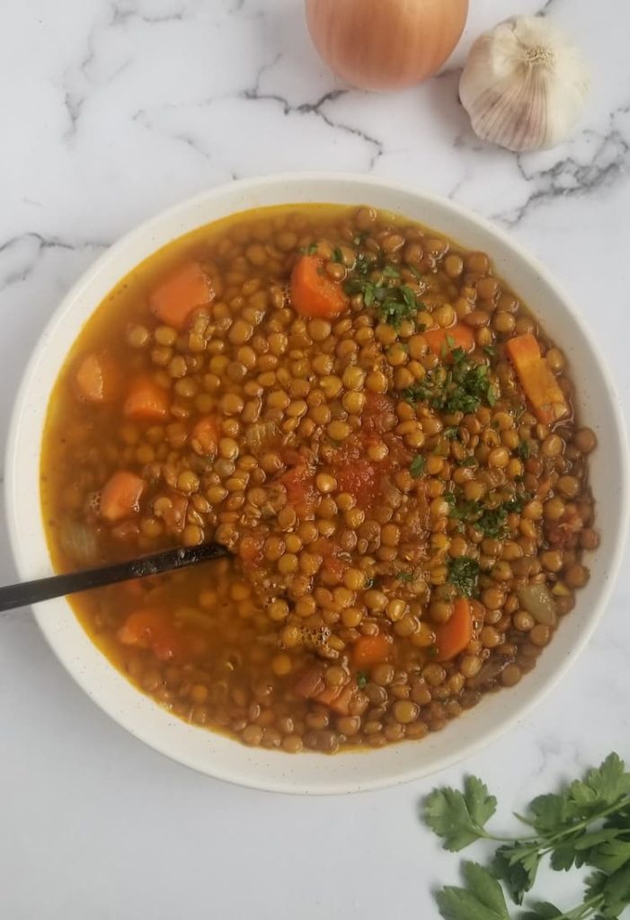 big bowl of lentil soup with carrots, tomatoes, onions and parsley, whole onion and garlic bulb in the background, fresh parsley on the bottom
