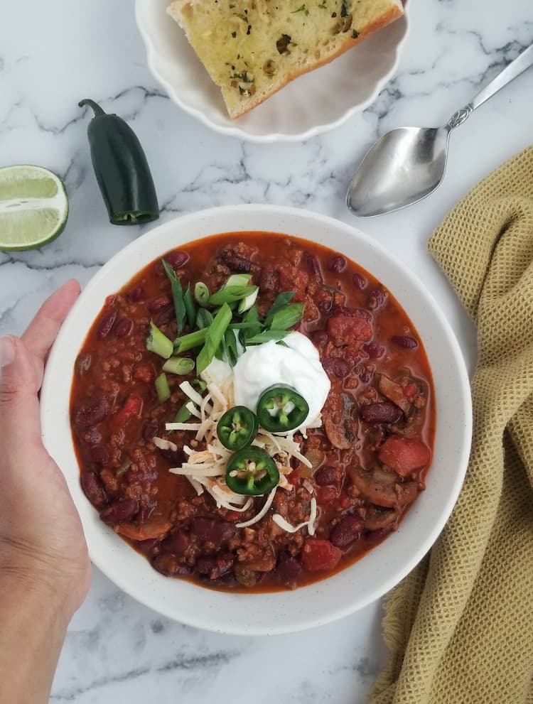 hand holding a big bowl of chili on one side, chili is topped with grated cheese, sliced jalapeno and green onions and sour cream, garlic bread, half a jalapeno and lime and a silver spoon in the background
