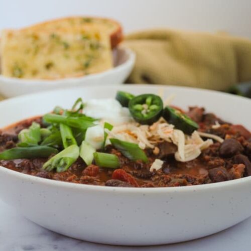 big bowl of chili topped sour cream, shredded white and oranges cheeses and sliced jalapeno and green onions, garlic bread, jalapeno and half a lime in the background