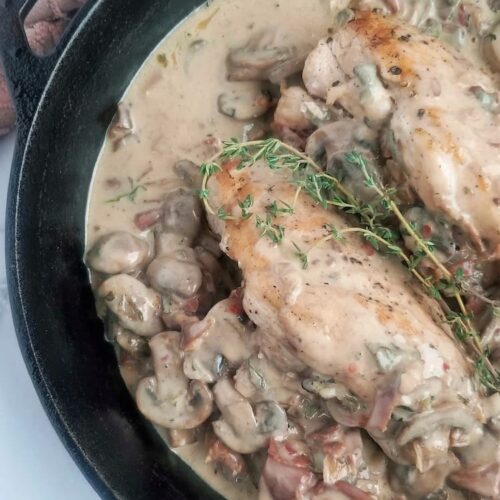 black cast iron skillet with two chicken breasts and creamy mushroom and prosciutto sauce, fresh thyme on top