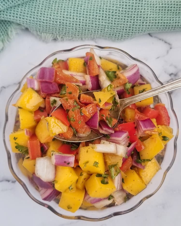 chunks of mango, red onion, parsley, red pepper and habaneros in a small glass bowl with a spoon in the middle holding some