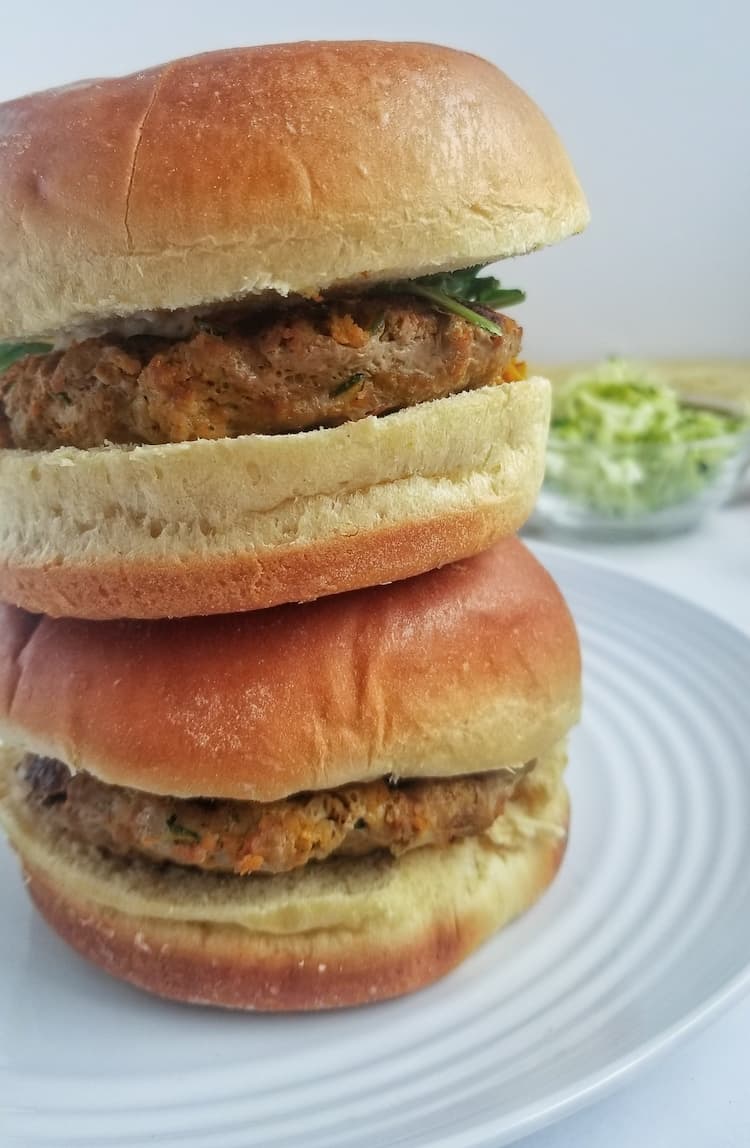 2 stacked zucchini turkey burgers on buns on a white plate, small bowl of shredded zucchini in the background