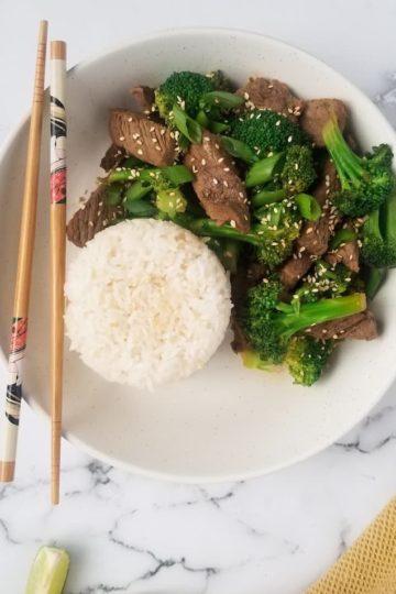 bowl of beef and broccoli, pile of rice in the bowl beside it, chopsticks on the bowl, 1 lime wedge at the bottom left of the picture