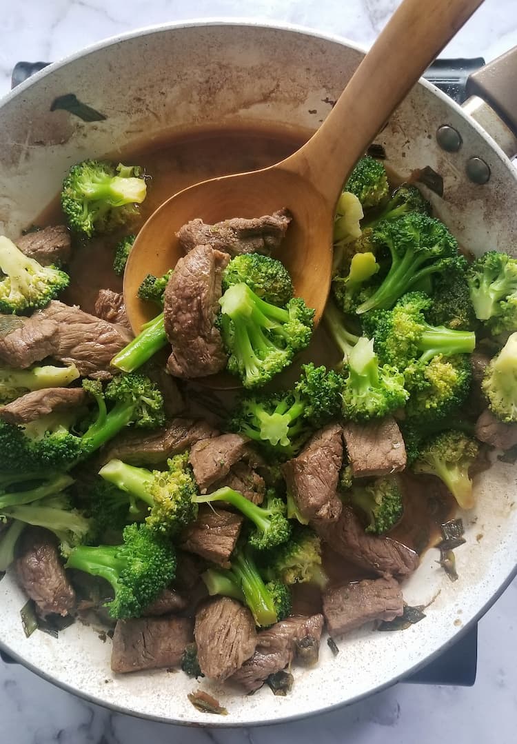 skillet with beef and broccoli in a brown sauce with a big wooden spoon and some of the beef and broccoli on the spoon