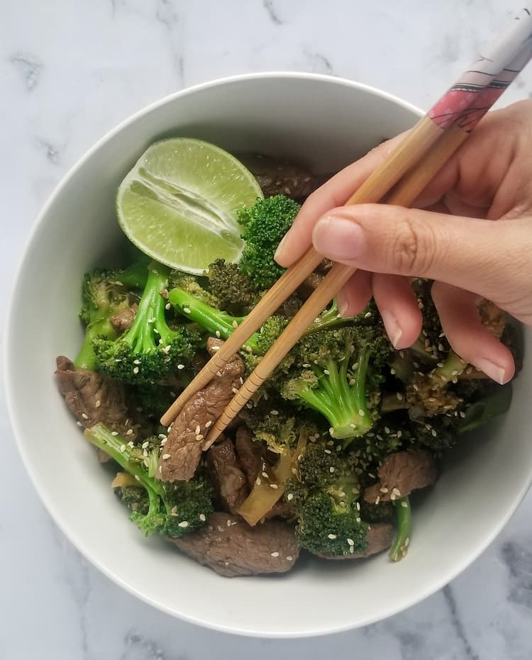 bowl of beef and broccoli with half a lime, hands with chopsticks holding a piece of beef