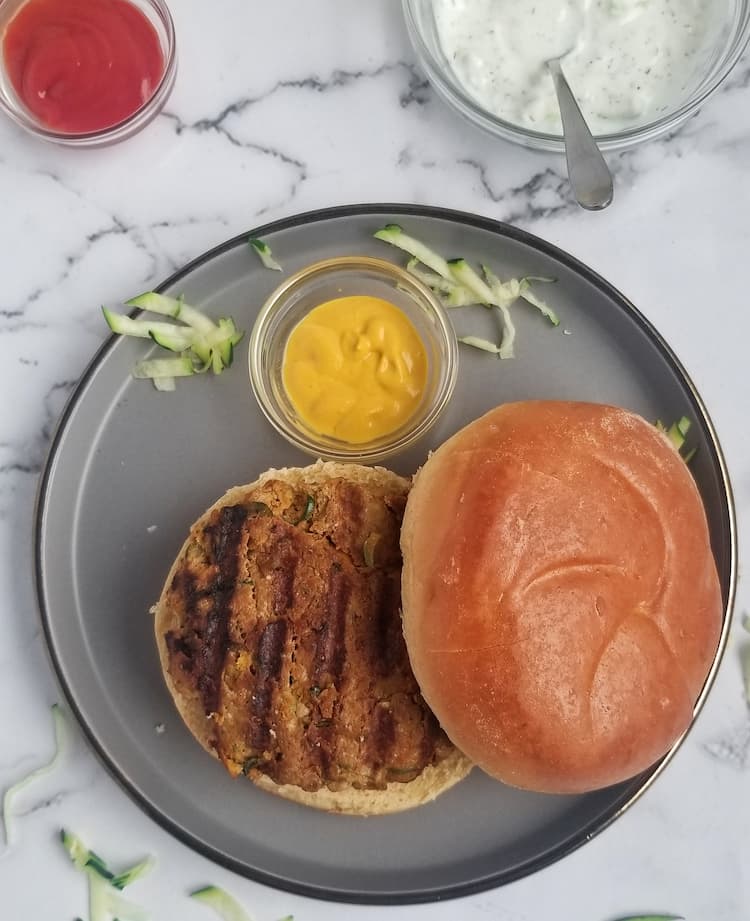 a grilled zucchini turkey burger on a bun on a plate with a small ramekin of mustard, ketchup and white dip in the background, shredded zucchini pieces around