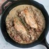 black cast iron skillet with two chicken breasts and creamy mushroom and prosciutto sauce, fresh thyme on top