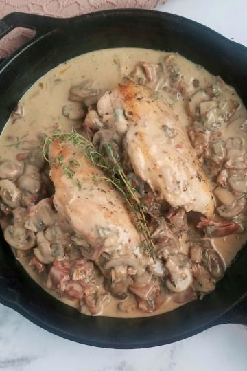 black cast iron skillet with two chicken breasts and creamy mushroom and prosciutto sauce, fresh thyme on top