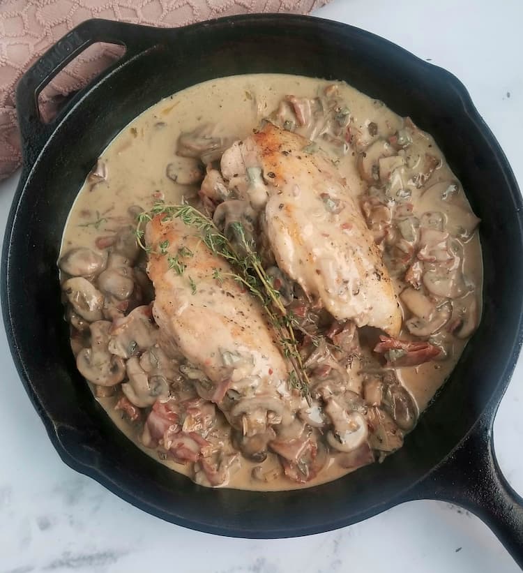 black cast iron skillet with two chicken breasts and creamy mushroom and prosciutto sauce, fresh thyme on top
