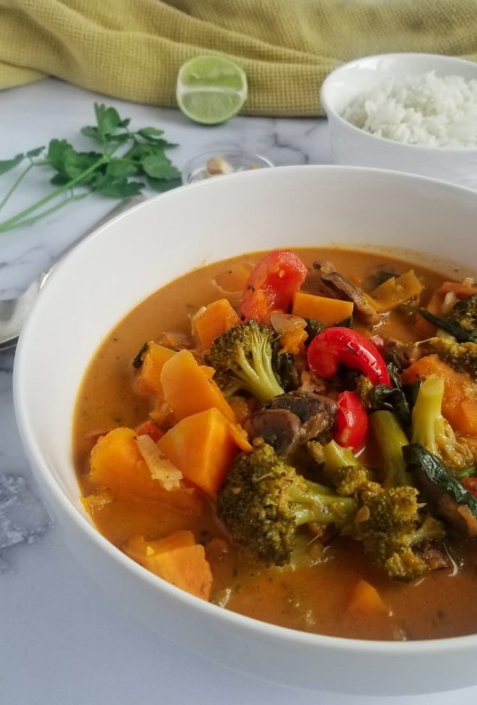 side angel of bowl of sweet potato curry with cubed sweet potatoes, red peppers, broccoli, mushrooms, orange broth, bowl of rice and half a lime and a bunch of parsley in the background