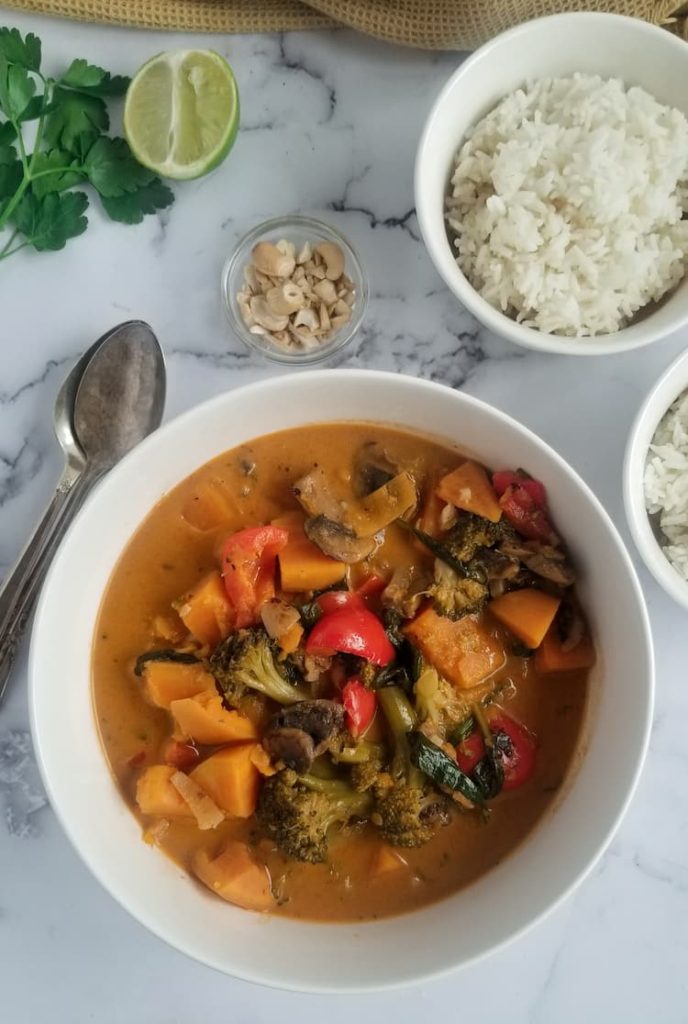 big bowl of sweet potato curry with veggies such as red pepper chunks, spinach and broccoli florets, bowl of rice and cashews in the background with a half lime and bunch of parsley