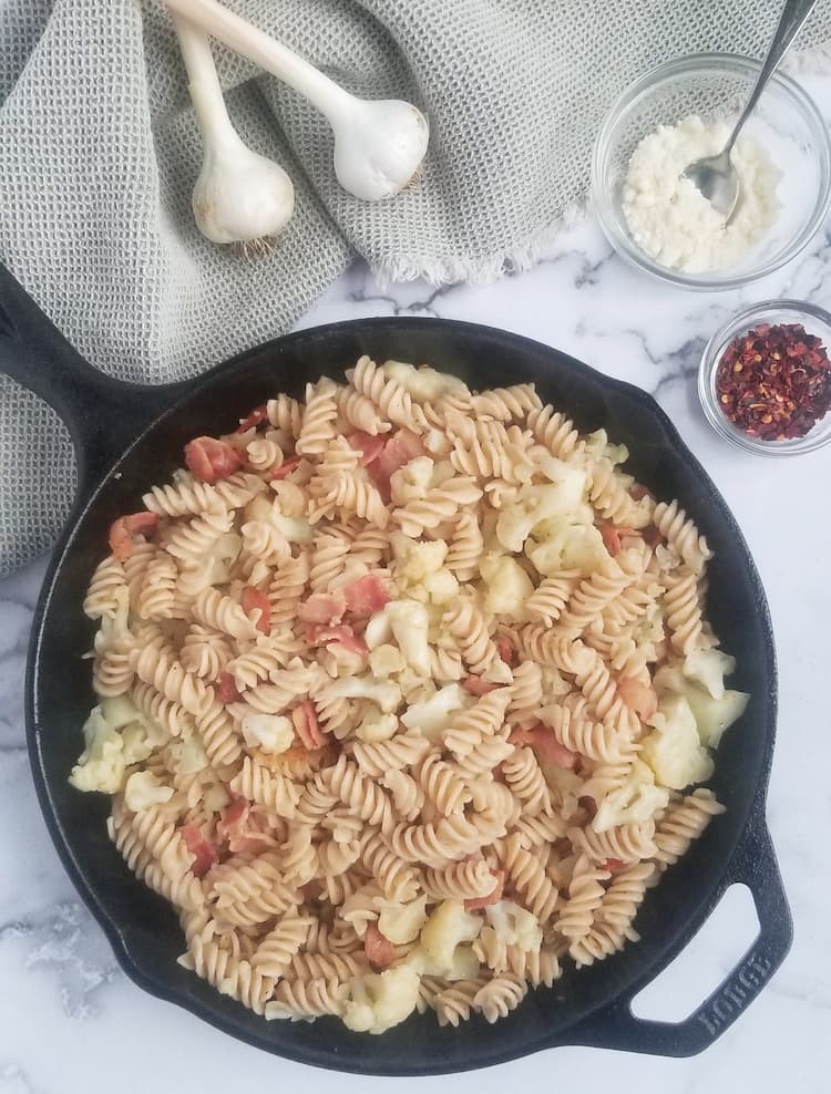 large black cast iron skillet of curly brown rice pasta with bacon and cauliflower pieces, two garlic bulbs in the background, a small clear bowl of parmesan cheese with a small silver spoon in it and a smaller bowl with chili flakes in it beside the skillet
