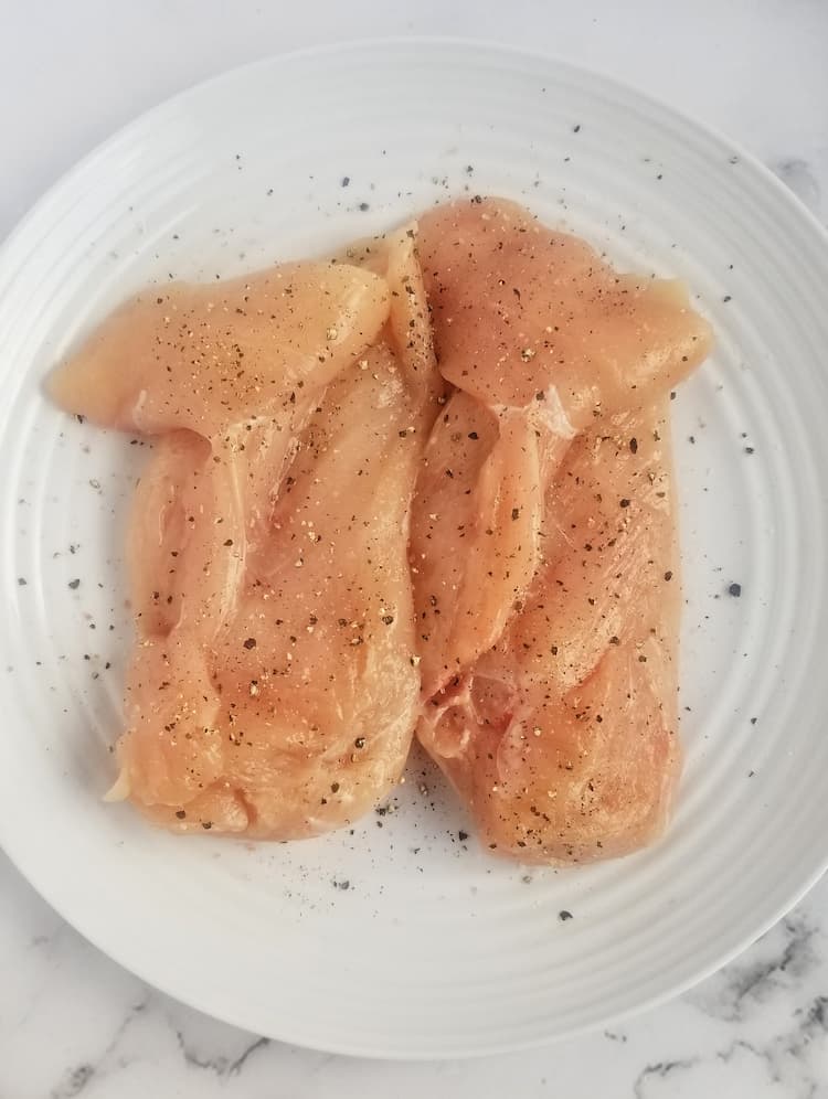 two raw chicken breasts on a plate seasoned with salt and pepper