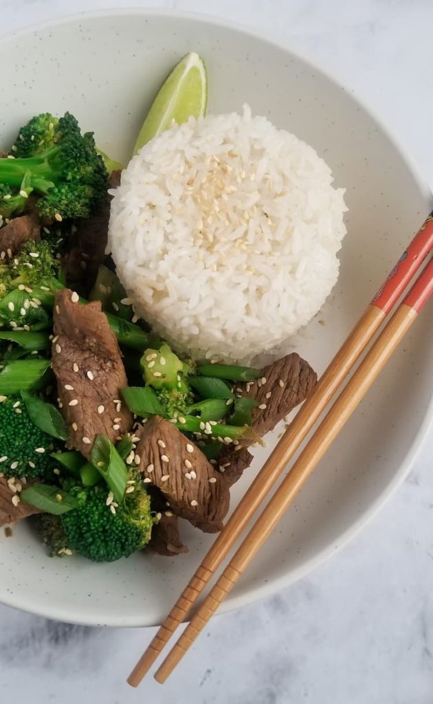 bowl of beef and broccoli, lime wedge, pile of rice and chopsticks in the bowl